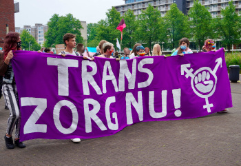 Foto van activisten (met mondkapjes) met die op straat lopen met een groot paars spandoek met de tekst "Trans Zorg Nu!". Op de achtergrond zijn bomen zichtbaar.