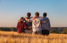 Four young people standing next to each other, in a row, their arms around each others shoulders. They stand with their backs to the camera, in a field of grass or grain, seemingly looking to the horizon. In the background we see a forest and a blue sky, without clouds.