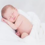 Een witte baby met rossig haar ligt met gesloten ogen, het hoofd op de armen en de benen onder zich gevouwen in een (grote) witte handddoek.