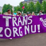 Foto van activisten (met mondkapjes) met die op straat lopen met een groot paars spandoek met de tekst "Trans Zorg Nu!". Op de achtergrond zijn bomen zichtbaar.