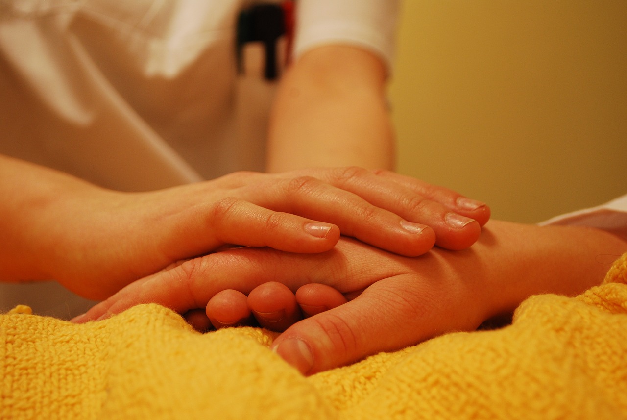 De beide handen van een iemand met een witte jas met korte mouwen (vermoedelijk een verpleegkundige) houdt een hand vast, die op een gele deken ligt.